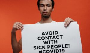 A serious-looking man stands against an orange background holding a large white sign that reads, “AVOID CONTACT WITH SICK PEOPLE! #COVID19” in bold black letters.